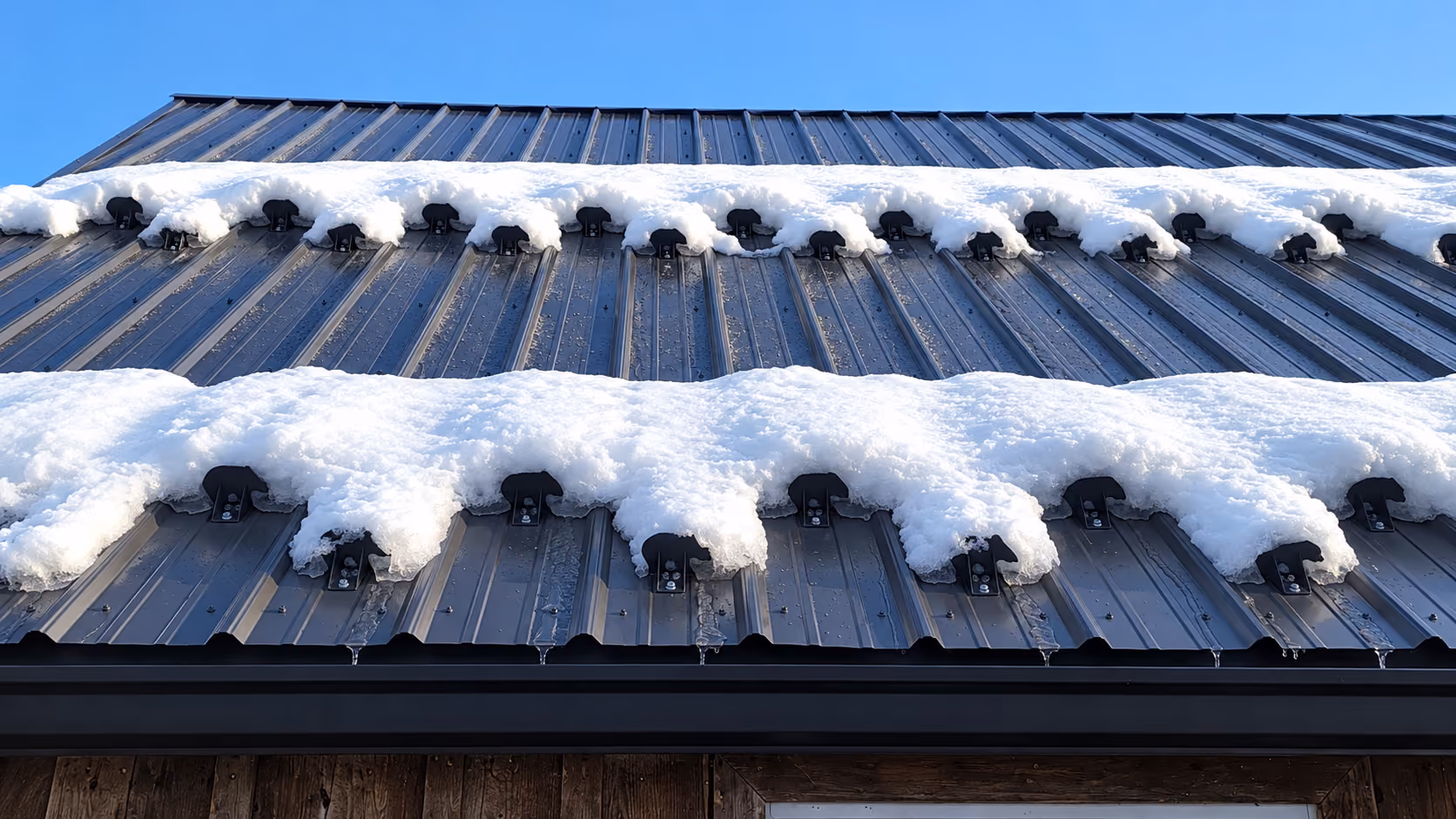 Two staggered rows of pad-style snow guards holding snow on a metal roof above the eave, Eagle River