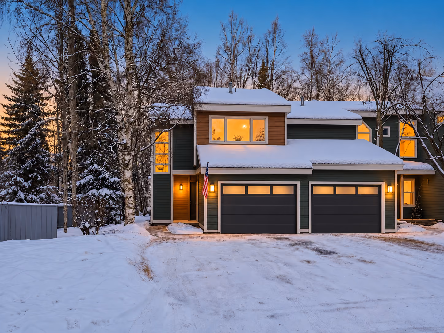 Two-story Anchorage home with snow on its low-slope roofs at winter dusk, birch and spruce around it